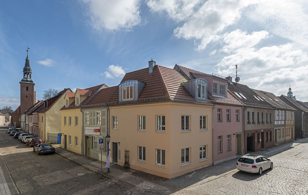 Blick auf die Häuserecke Heiligegeisttraße und Am Rosenwinkel. Im Vordergrund das lachsfarbene Haus Heiligegeisttr. 18, links davon Haus Nummer 16 in grau und rechts Am Rosenwinkel 15 in rot. Alle Häuser haben Schrägdächer aus roten Ziegeln.